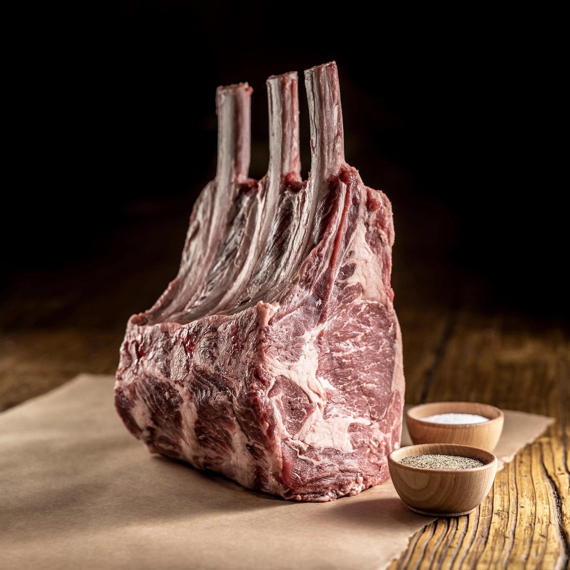 uncooked 3 bone tomahawk ribeye on parchment paper with cups of seasoning